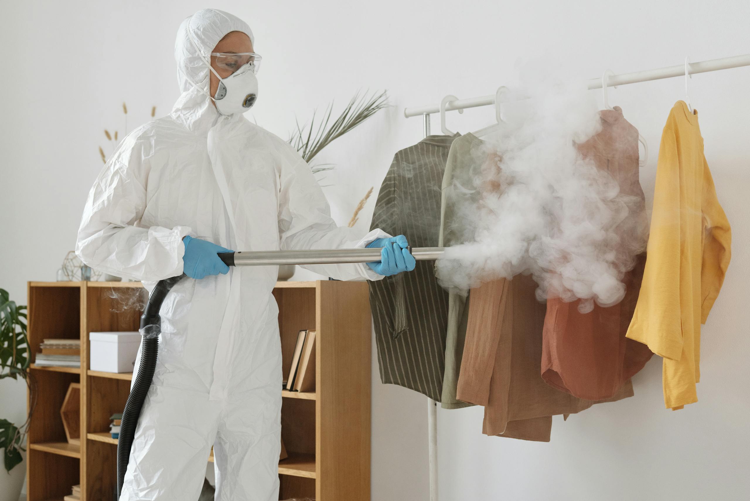 A person in PPE suit using a fumigation device to disinfect clothes indoors.